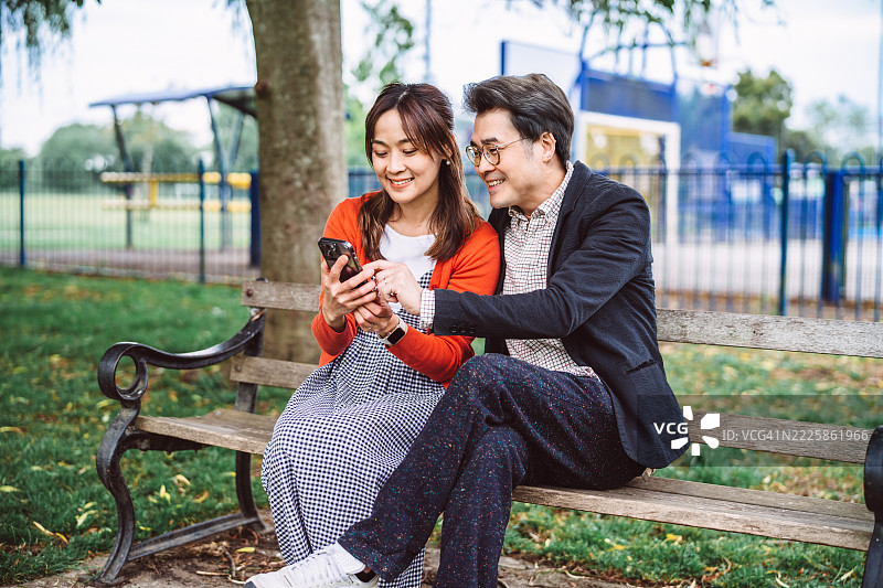 快乐的情侣在公园长椅上一起使用智能手机图片素材