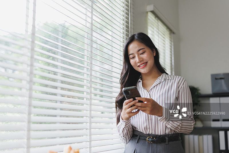 年轻的亚洲女商人在办公室窗边使用手机图片素材