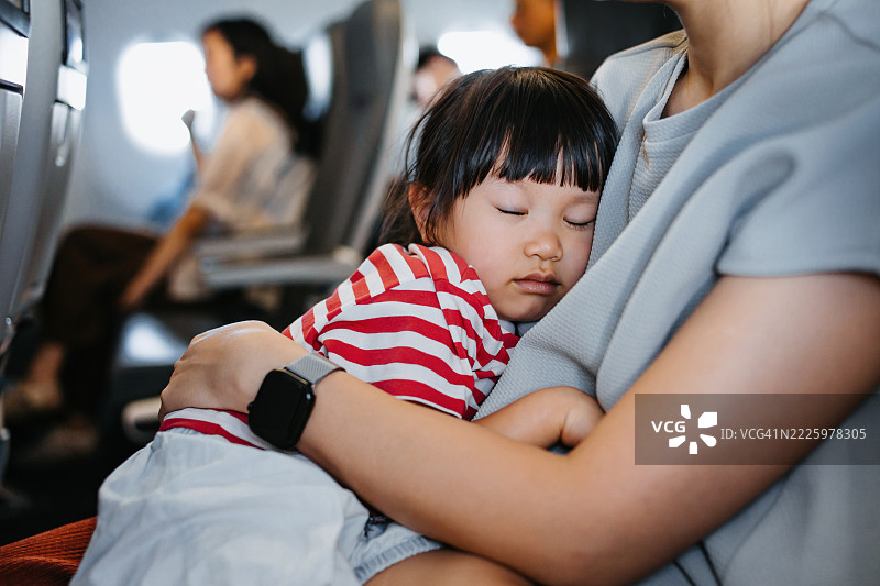 小亚洲女孩在飞机上舒适地在母亲怀里小睡，和家人一起旅行。家庭生活方式。家庭旅行和度假。图片素材