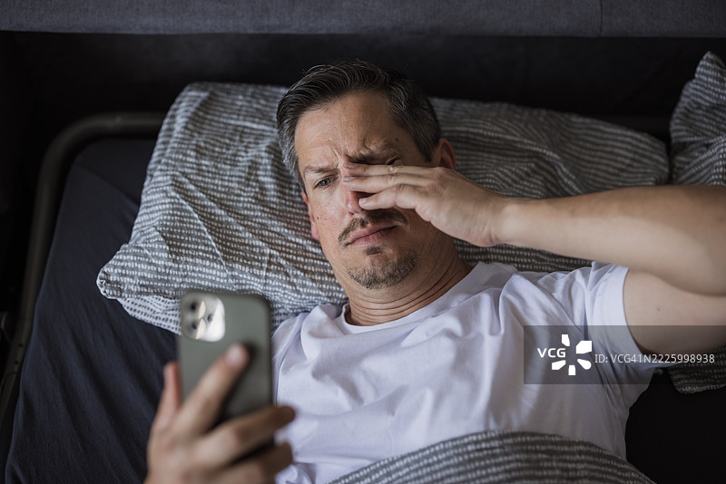 疲惫的男人在床上使用智能手机时揉眼睛图片素材
