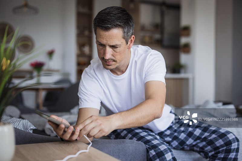 穿着睡衣的男人在家里给智能手机充电图片素材