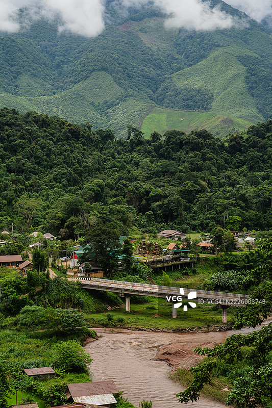 一个被茂密绿色森林和山丘环绕的乡村，拥有一座跨越泥泞河流的混凝土桥，天空阴云密布。图片素材