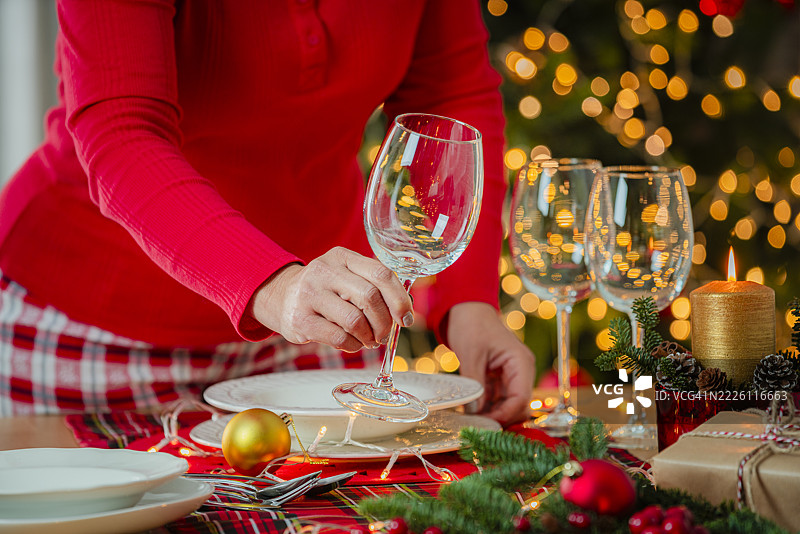 女人为圣诞晚餐、跨年夜或餐桌布置摆放餐具。酒杯和装饰蜡烛。点亮的圣诞树。图片素材