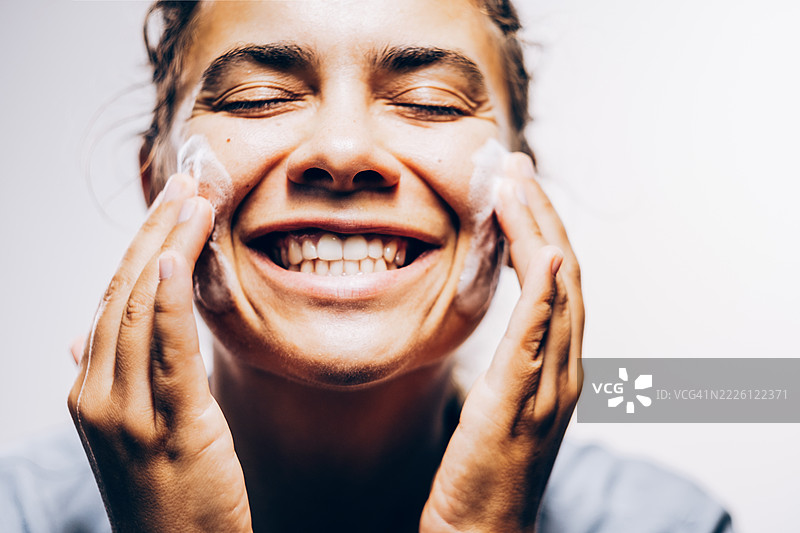 微笑的美丽女性用双手将泡沫洁面乳涂抹在脸颊上。工作室特写，背景为白色。图片素材