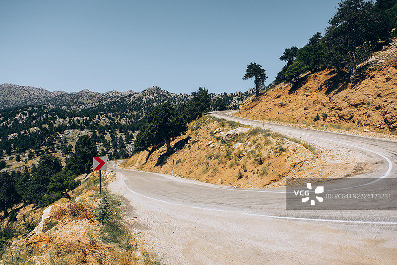 蜿蜒的沥青公路在晴朗的蓝天下穿过山区，稀疏的植被和松树沿着山坡生长。图片素材