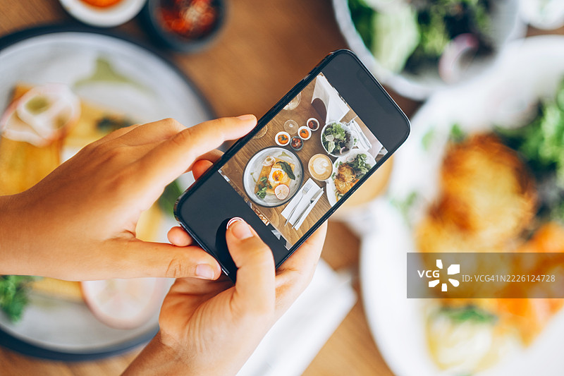 手握智能手机拍摄早餐桌的照片。周围有多道菜肴和饮品。美食摄影和社交媒体分享的概念。图片素材