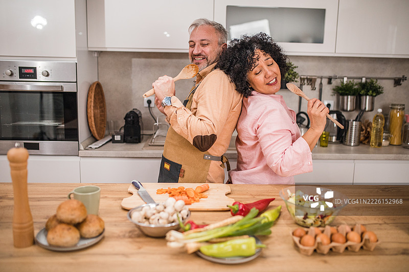 一对多民族情侣在温馨的厨房里准备午餐，享受着乐趣。他们一边唱歌一边笑，使用木勺当作假麦克风，把日常烹饪变成充满爱与玩乐的快乐表演。图片素材