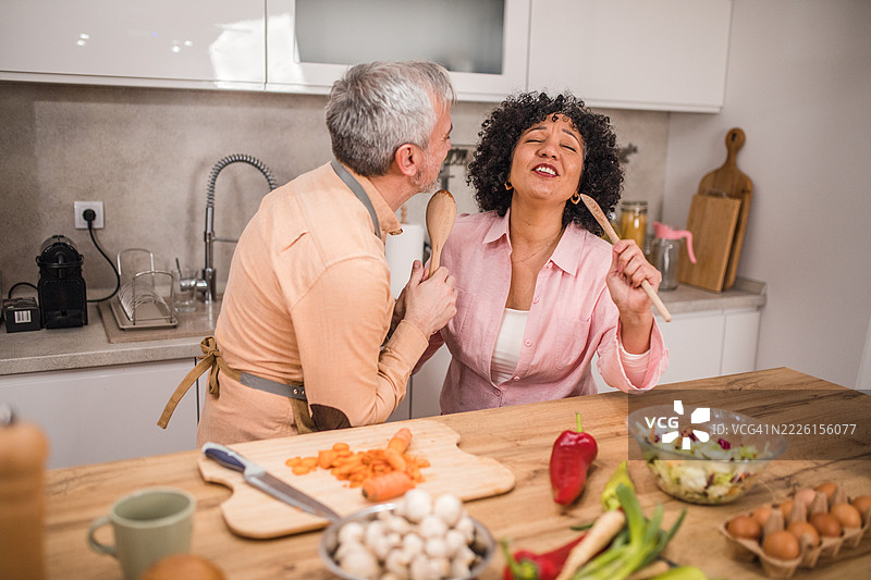 一对多元化的情侣在家享受有趣的烹饪时光，把厨房变成了一个小舞台。他们像麦克风一样握着木勺，一边唱歌一边跳舞，共同准备健康的餐点。图片素材