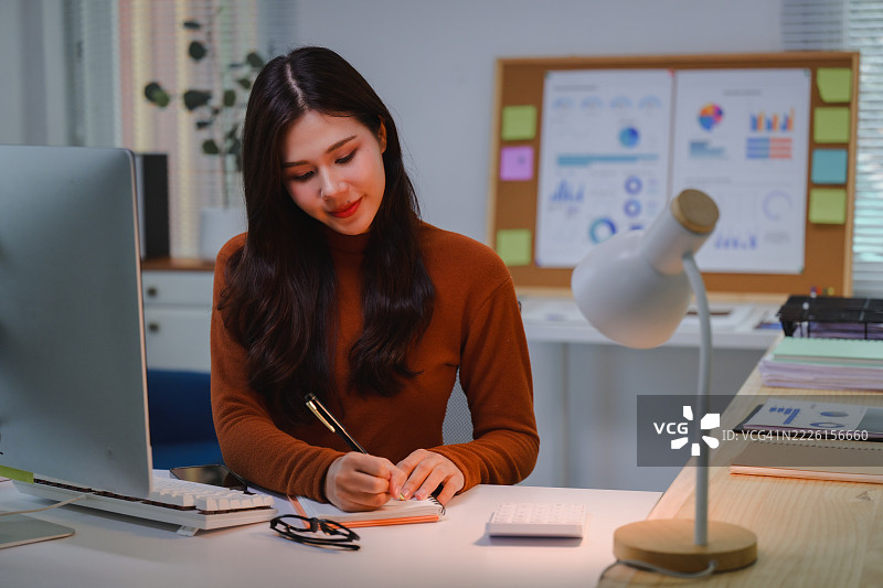年轻的亚洲女商人在家办公，正在笔记本上写笔记，桌上有电脑和文件。图片素材
