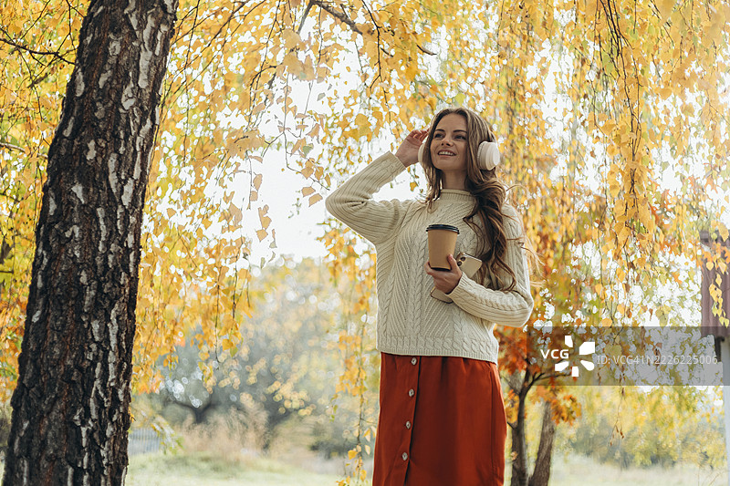 微笑的年轻女性在黄桦树下戴着耳机听音乐，同时手里拿着一次性杯子和智能手机。图片素材