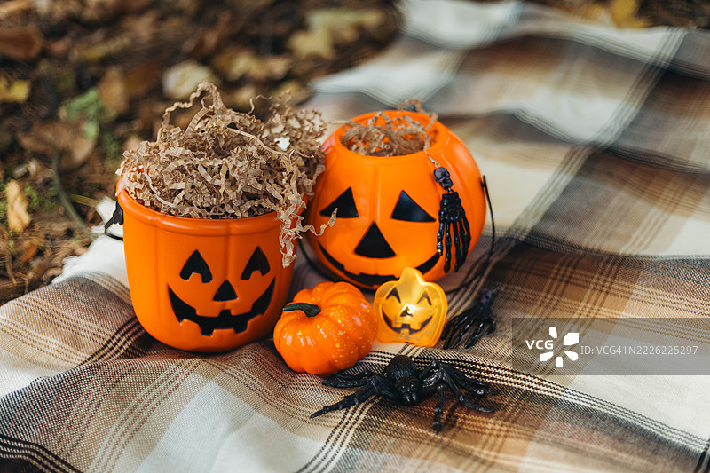 格子毛毯上放着两个装满皱纸的节日橙色南瓜形状的桶，一个发光的南瓜蜡烛和一只黑色蜘蛛。图片素材