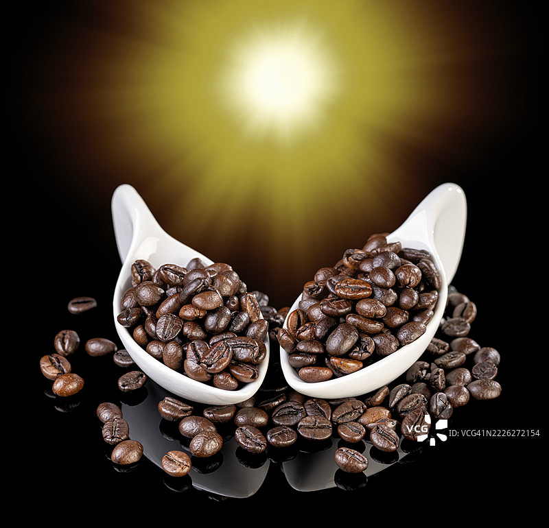 光滑黑色背景上，阳光照射下的两只瓷勺内盛放着烘焙咖啡豆。图片素材