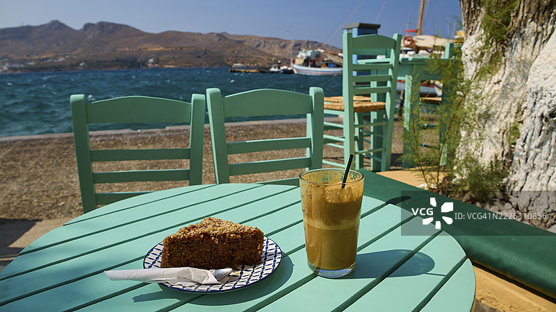 海边阳光明媚的放松氛围中，蓝绿色的桌子上摆放着饮料和蛋糕，地点位于希腊多德卡尼斯群岛的莱罗斯岛阿吉亚玛丽娜。图片素材