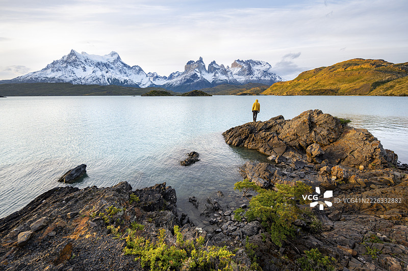 傍晚时分，年轻男子站在佩霍湖（Lago Pehoe）岸边，背景是百内圆锥山（Cuernos del Paine），位于百内国家公园（Torres del Paine National Park），智利。图片素材