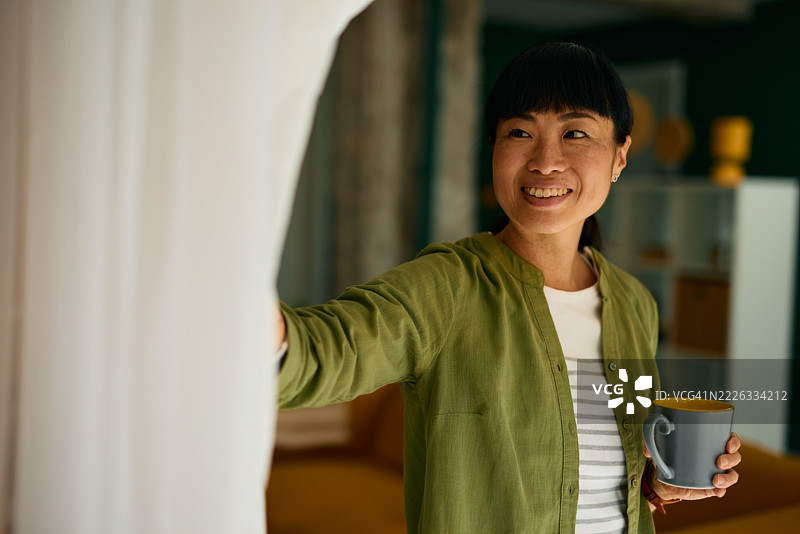 微笑的女人拉开窗帘，手里拿着咖啡杯图片素材
