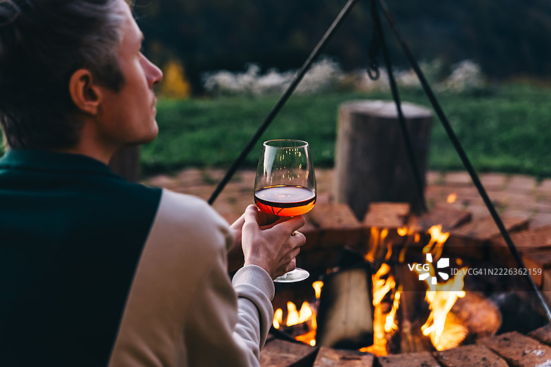 男人在日落时分，坐在火边享受山区秋季风景，手中拿着一杯葡萄酒。图片素材