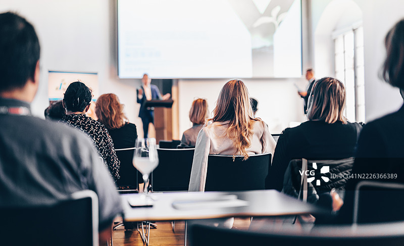 在商业活动的会议厅中，演讲者正在发言，观众的背影不可辨认。图片素材
