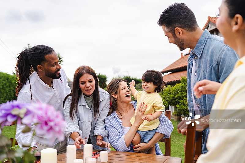 多元文化家庭聚会，父母在阳光明媚的后院庆祝活动中与微笑的幼儿玩耍，共享快乐时光。图片素材