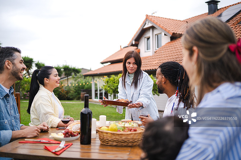 多元化的朋友们在后院共享美餐，一位女性在温馨的木桌上呈现烤鸡，气氛融洽。图片素材