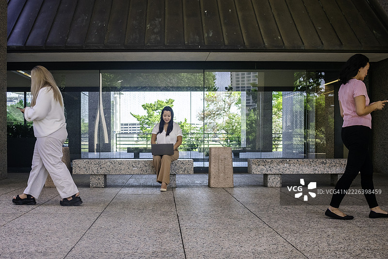 一位女商人坐在长椅上使用笔记本电脑，旁边两位女性一边走过一边使用智能手机。图片素材