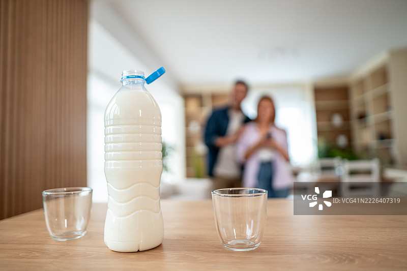 厨房桌子上有新鲜牛奶瓶和空玻璃杯，背景中有一对情侣。图片素材