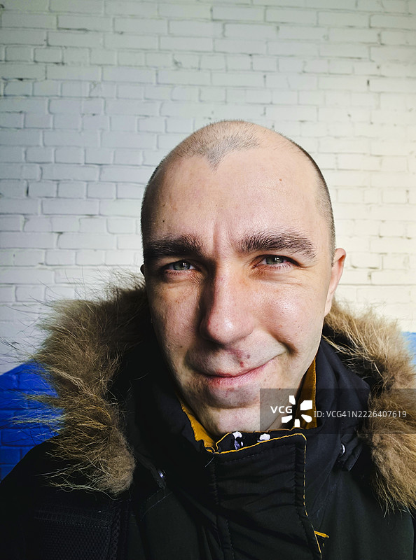 微笑的男子，光头明显，穿着毛皮衬里的夹克，站在有纹理的砖墙前，在这一瞬间散发出温暖和个性。图片素材