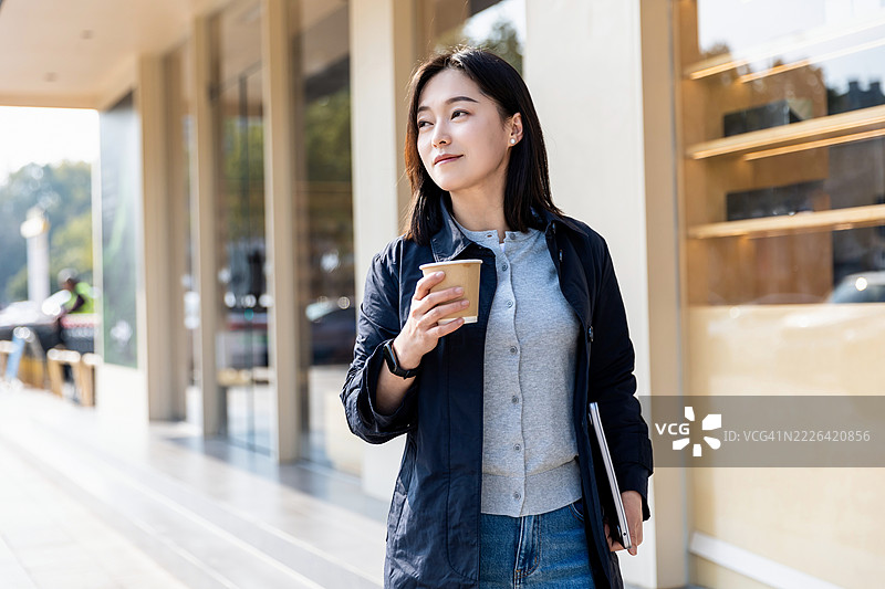 快乐成功的亚洲女商人手持外卖咖啡杯和笔记本电脑，站在玻璃建筑旁的街道上图片素材