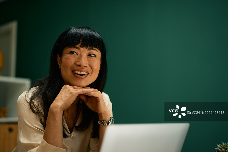 微笑的女商人一边工作一边看向远方，手边放着笔记本电脑图片素材