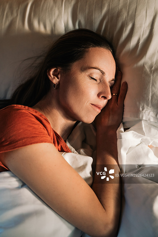 一位美丽的年轻女性在酒店的白色床上睡觉，窗外的光线洒在她的脸上。图片素材