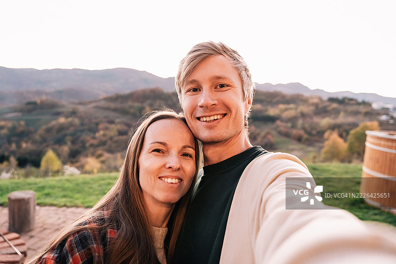 微笑的情侣在户外拍摄自拍，背景是金色秋天的山丘和落日的天空，捕捉到一起快乐的旅行时刻。图片素材