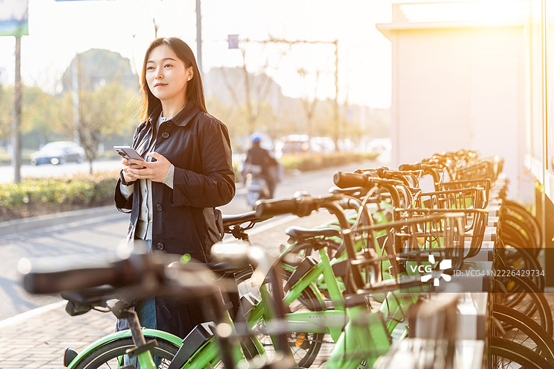 年轻亚洲女性在城市中使用租赁自行车，手持智能手机。共享交通服务。科技与可持续生活理念的生活方式。图片素材