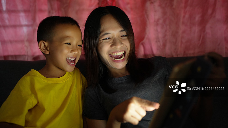 快乐的女人和孩子在柔和的光线下共享欢笑时刻，目光投向平板电脑。他们的表情展现了幸福和亲密感。图片素材