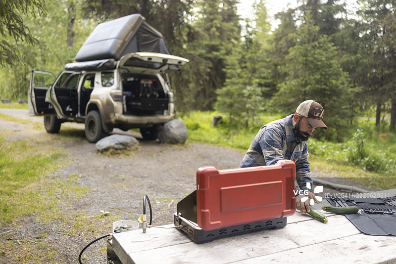 一名男子在露营旅行中使用便携式露营炉准备餐食，背景是一辆配备越野露营或车顶帐篷的车辆。图片素材