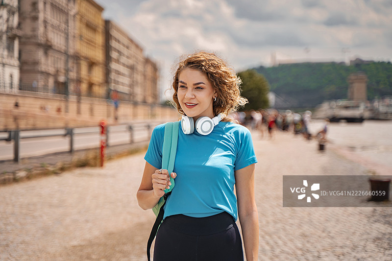 年轻的运动女性在多瑙河畔的布达佩斯走路，耳朵里戴着耳机，背着背包。图片素材