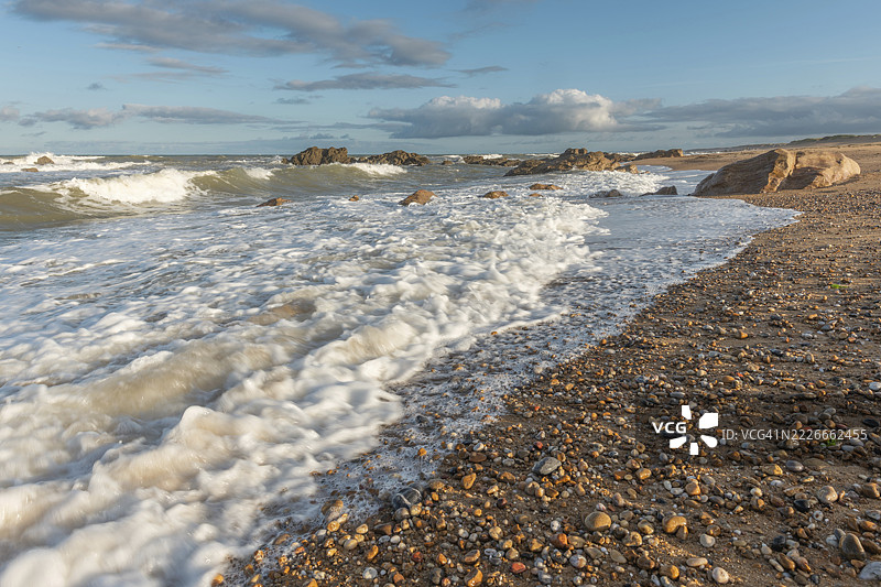 夕阳下，海浪轻轻拍打着岩石海岸，营造出宁静的氛围。沙滩上散落着卵石，四周环绕着美丽的天空。法国旺代省莱萨布尔多隆的奥布雷海滩。图片素材