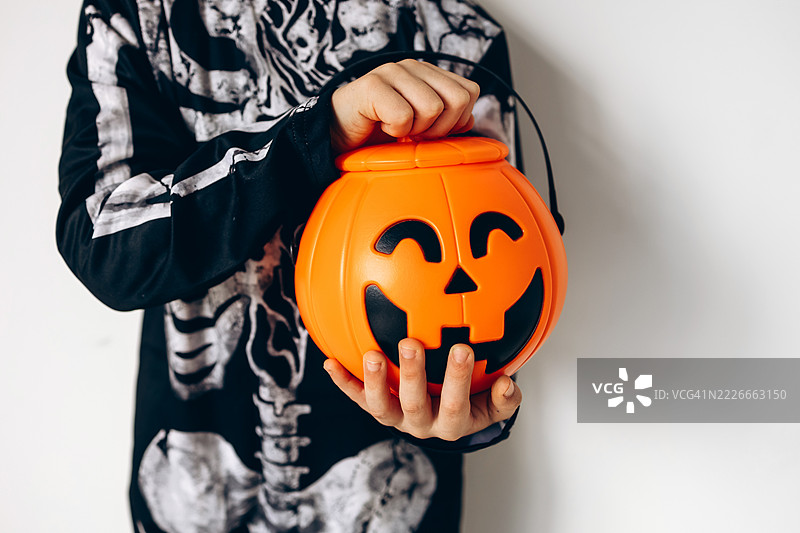 可爱男孩穿着黑色骷髅服，手里拿着南瓜形状的桶，背景为白色。图片素材