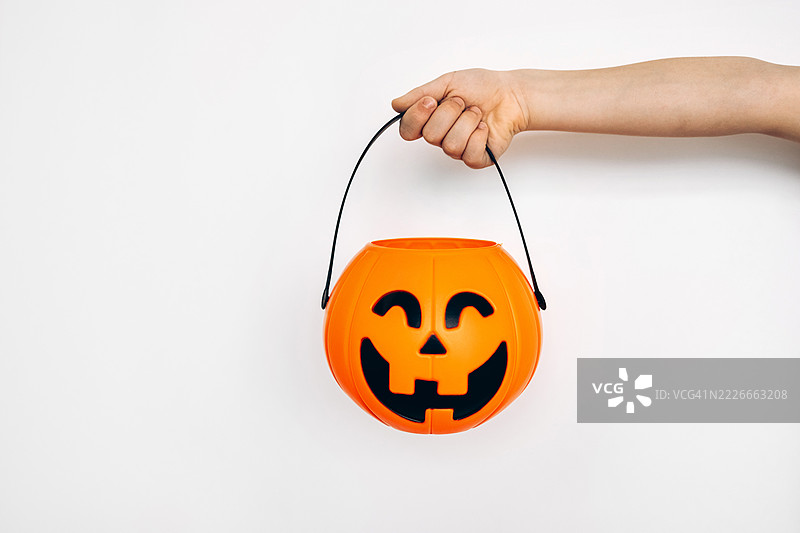 孩子的手握着万圣节南瓜糖果桶的特写，背景为白色。快乐的万圣节概念图片素材