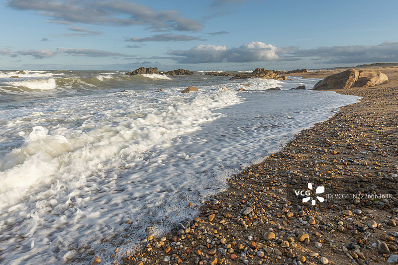 夕阳下，海浪轻轻拍打着岩石海岸，营造出宁静的氛围。沙滩上散落着卵石，四周环绕着美丽的天空。法国旺代省奥朗海滩。图片素材