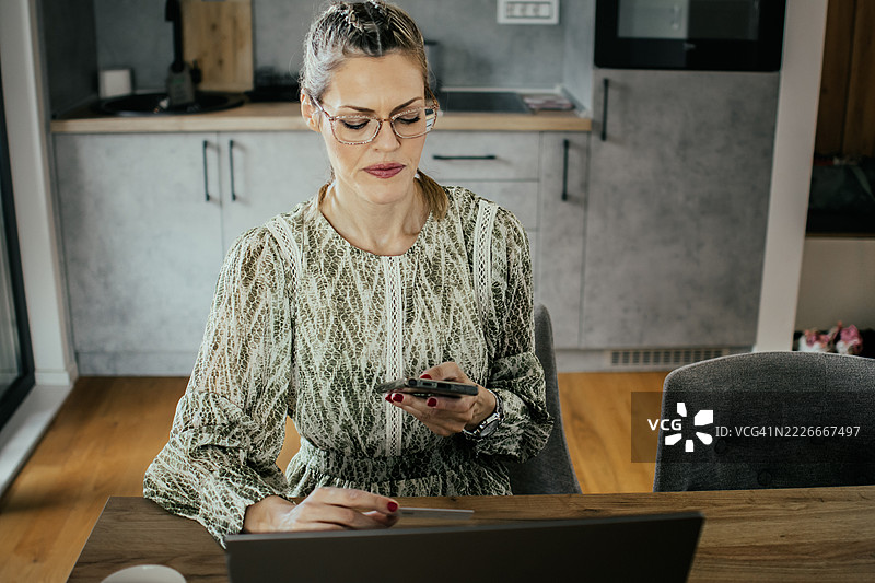 金发女子戴着眼镜，在现代公寓里使用笔记本电脑和银行信用卡进行网上购物，为家人购买礼物并支付账单。图片素材