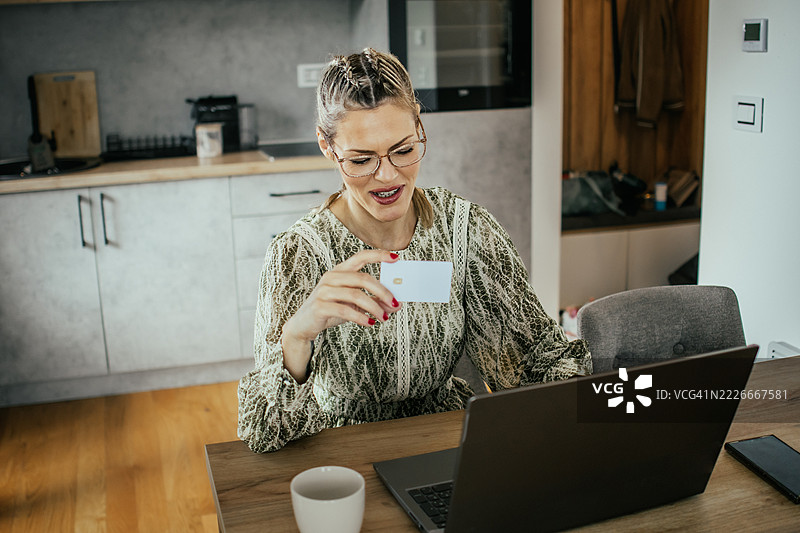 金发女士戴着眼镜，在现代公寓使用笔记本电脑，刷银行信用卡进行在线购物，为家人购买礼物并支付账单。图片素材
