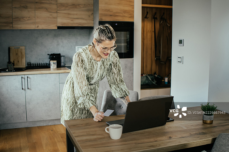 金发女子戴着眼镜，在现代公寓的笔记本电脑上使用银行信用卡进行在线购物，为家人购买礼物并支付账单。图片素材