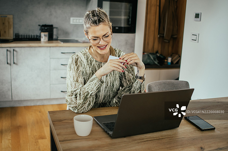 戴眼镜的金发女子在现代公寓的笔记本电脑上使用银行信用卡进行在线购物，为家人购买礼物并支付账单。图片素材