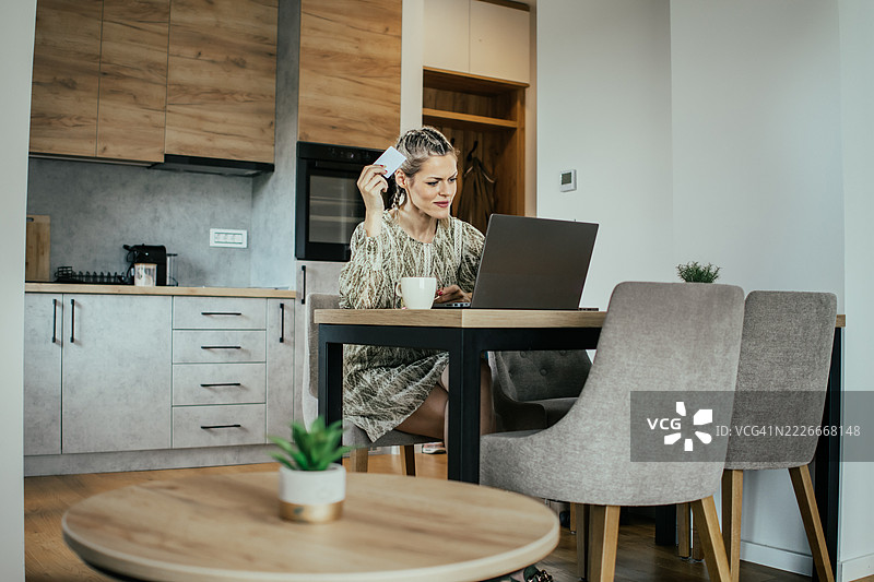 金发女子戴着眼镜，在现代公寓中使用笔记本电脑和银行信用卡进行在线购物，为家人购买礼物并支付账单。图片素材