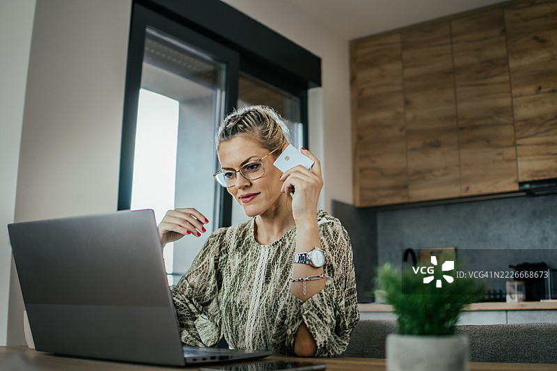 戴眼镜的金发女子在现代公寓的笔记本电脑上使用银行信用卡进行在线购物，为家人购买礼物并支付账单。图片素材
