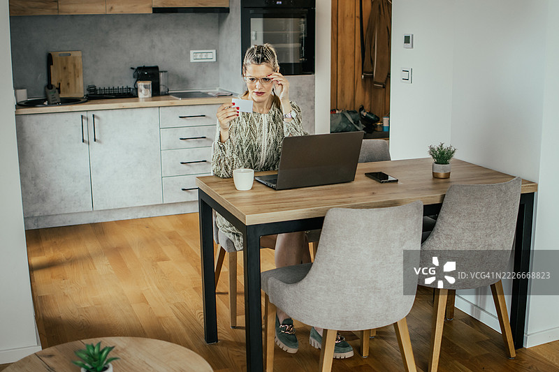 金发女子戴着眼镜，在现代公寓里使用笔记本电脑和银行信用卡进行在线购物，为家人购买礼物并支付账单。图片素材
