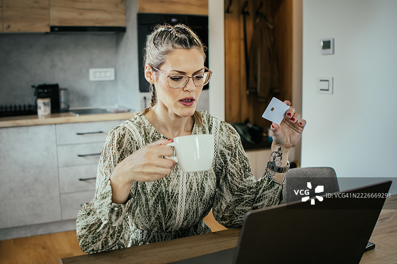 金发女士戴着眼镜，在现代公寓的笔记本电脑上使用银行信用卡进行在线购物，为家人购买礼物并支付账单。图片素材