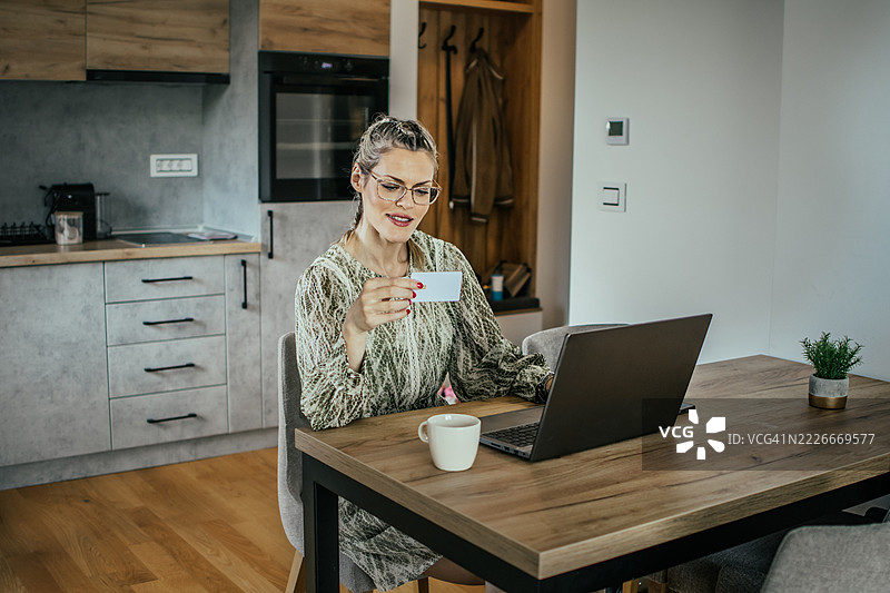 金发女士戴着眼镜，使用银行信用卡在现代公寓的笔记本电脑上进行网上购物，为家人购买礼物并支付账单。图片素材
