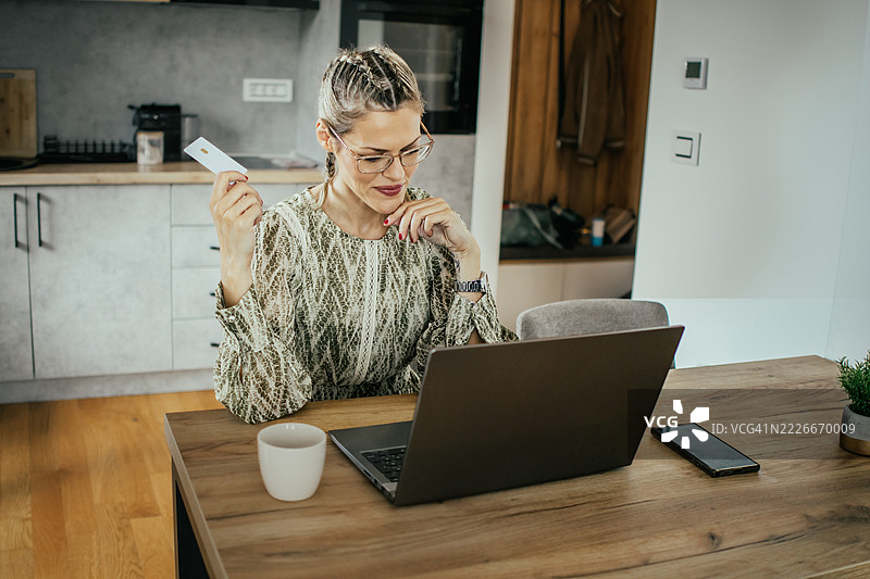 金发女士戴着眼镜，在现代公寓里用笔记本电脑使用银行信用卡进行在线购物，为家人购买礼物并支付账单。图片素材