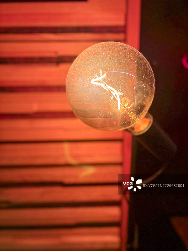 一盏发光的复古爱迪生灯泡挂在木条墙上，散发出温暖的橙色光芒。图片素材
