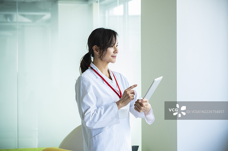 身穿白大褂的日本女医生在医院窗边思考地看着数字平板电脑图片素材